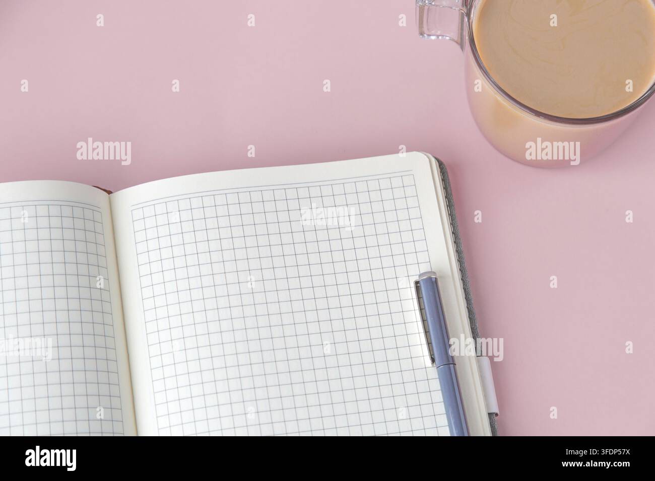 Ein offenes Gitter-Notizbuch mit einem Stift und einer Tasse Kaffee, die auf einem rosa Hintergrund platziert sind. Das minimalistische Arbeitsplatz-Setup repräsentiert Fokus und Produktivität. Co Stockfoto
