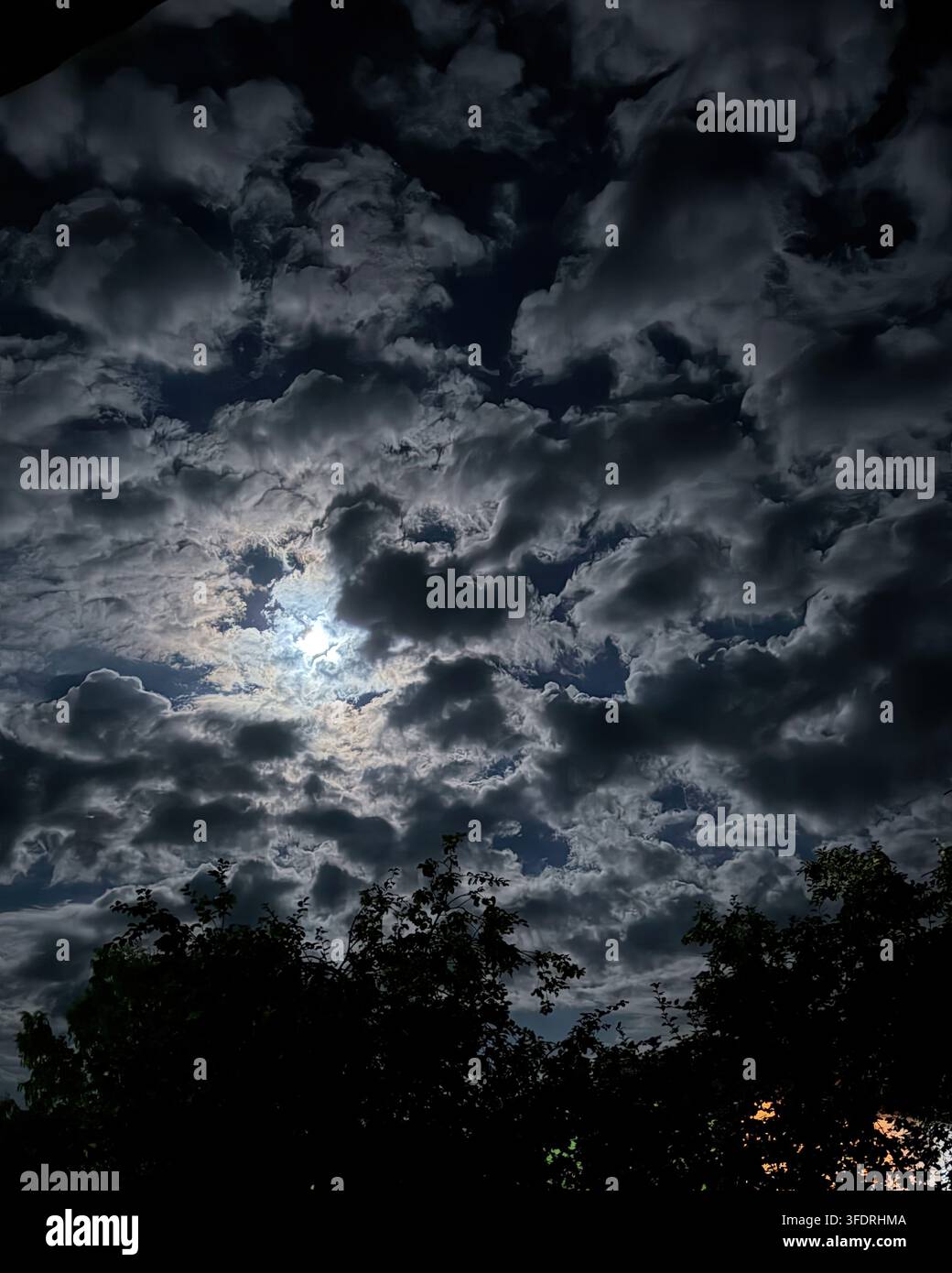 Dramatischer Nachthimmel mit hellem Mondlicht, das durch dunkle Wolken über Baumsilhouetten scheint Stockfoto