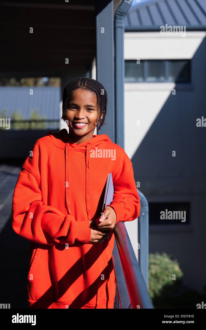 Afroamerikanischer Teenager mit orangefarbenem Kapuzenpullover, der sich auf einem Metallgeländer auf dem Balkon lehnt und einen dünnen Ordner hält Stockfoto