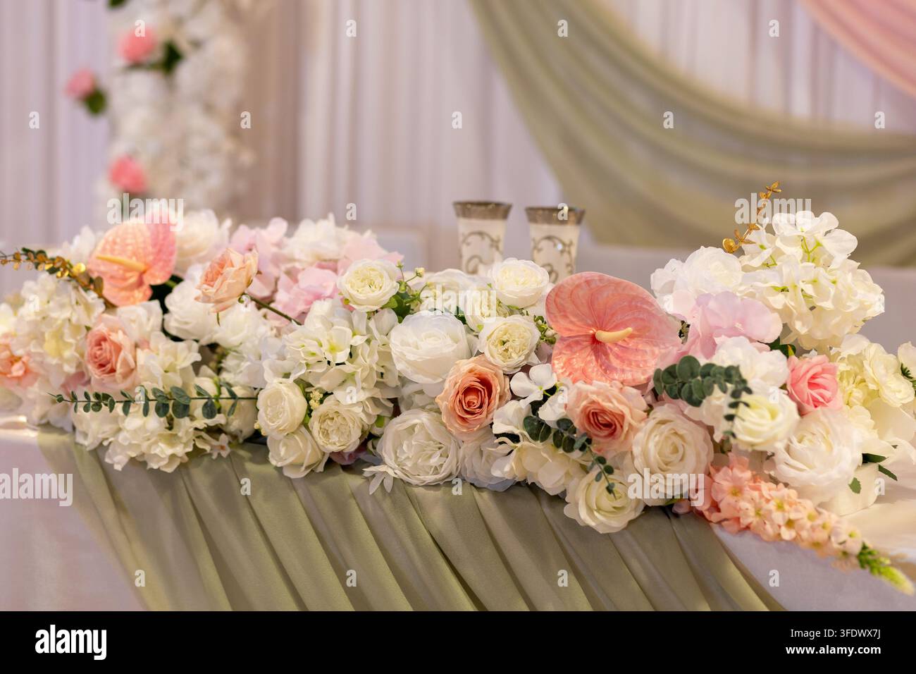 Ein Tisch ist mit einer Vielzahl von Blumen, darunter Rosen und Orchideen, für eine Hochzeitszeremonie gedeckt. Der Saal hat sanfte Beleuchtung und drapierten Stoffschmuck. Stockfoto