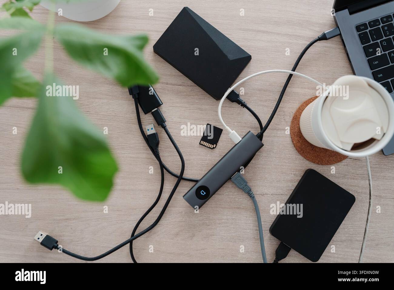 Verwirrte USB-Kabel, ein Dockingshub, externe Festplatten und eine Speicherkarte überladen einen Holzschreibtisch neben einem Laptop und einer Kaffeetasse. Unordentlicher Arbeitsplatz für Stockfoto