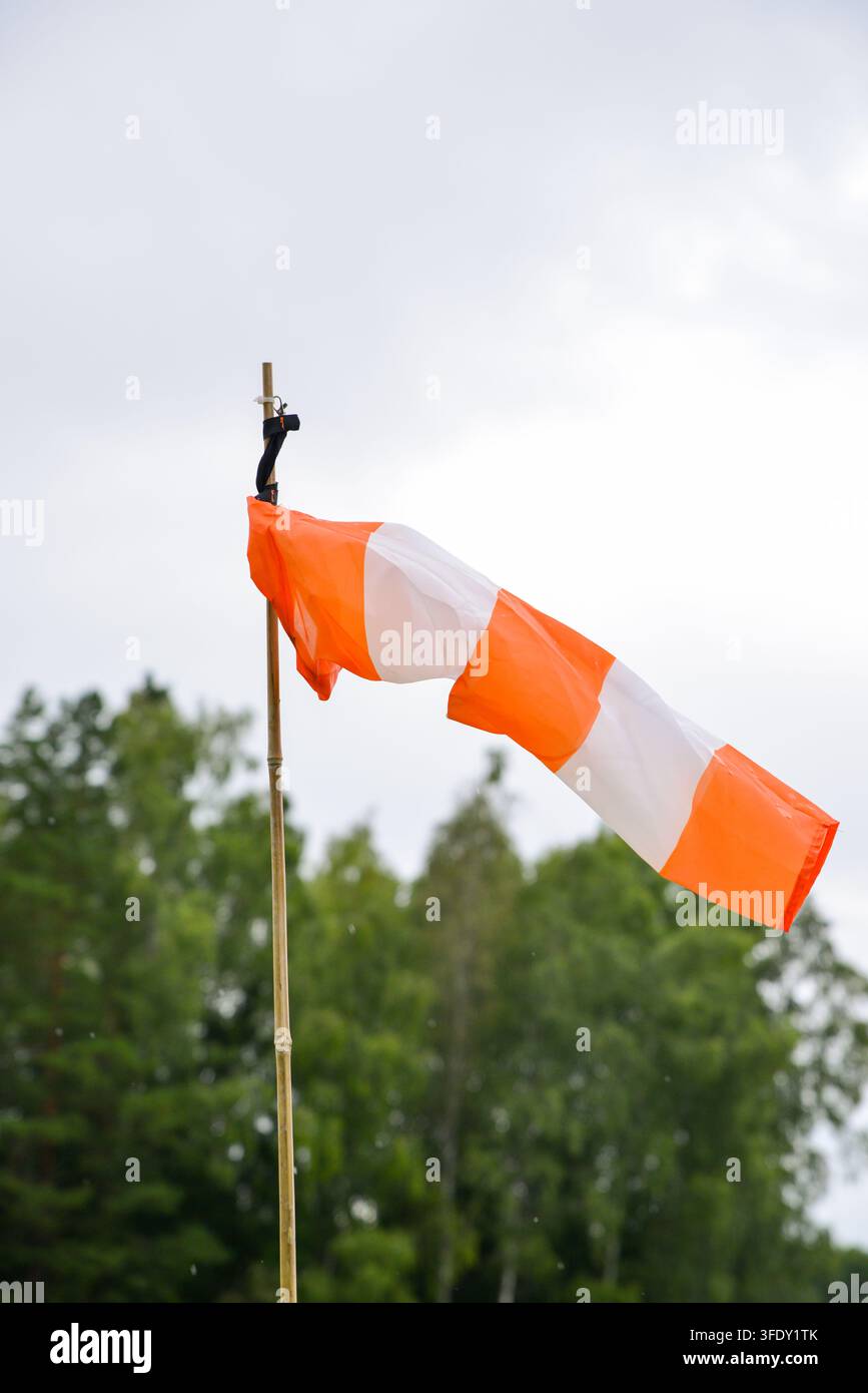 Helle orangefarbene und weiße Windsocke, die von einer gleichmäßigen Brise gegen einen weichen Himmel und grüne Bäume gespannt ist, veranschaulicht Windrichtung, Wetterbedingungen, hinaus Stockfoto