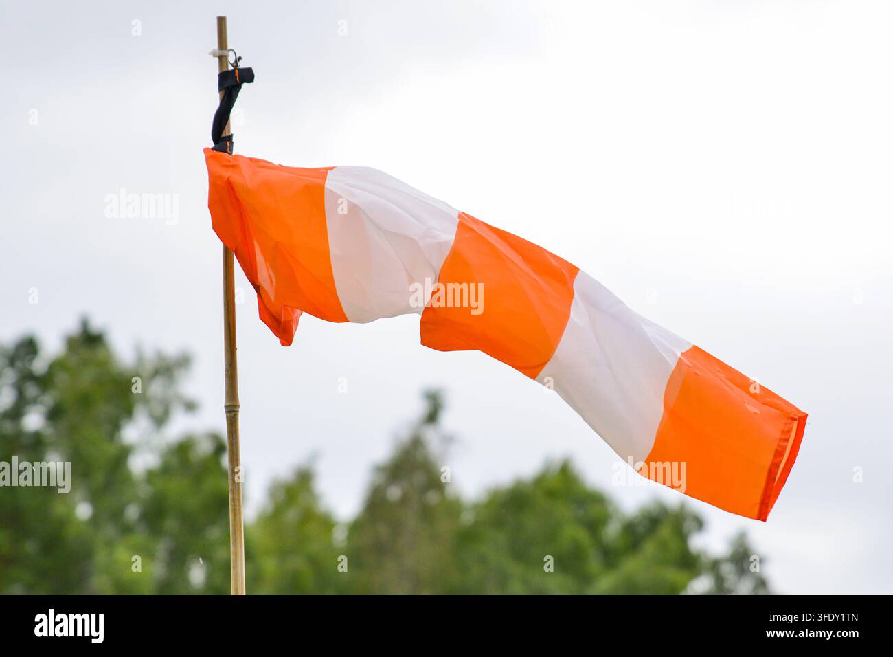 Helle orange-weiße Windsocke, die vom Wind auf einem Holzpfahl gegen einen hellen Himmel und verschwommene grüne Bäume gespannt ist, veranschaulicht die Windrichtung, wea Stockfoto