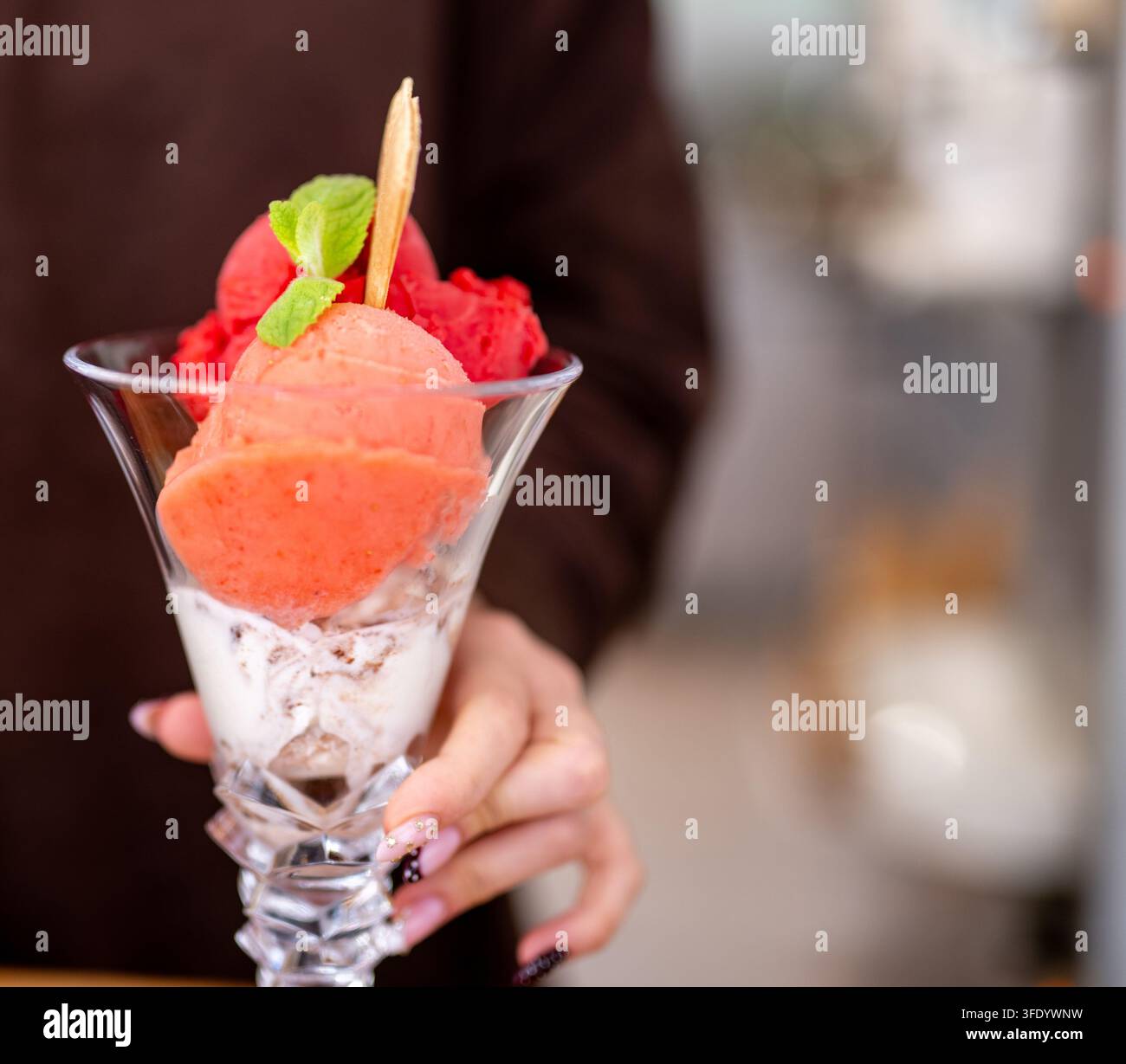 Hand präsentiert ein geschichtetes Dessert. Sahne, Pfirsich und rote Sorbet-Sphären füllen ein hohes Glas. Stockfoto