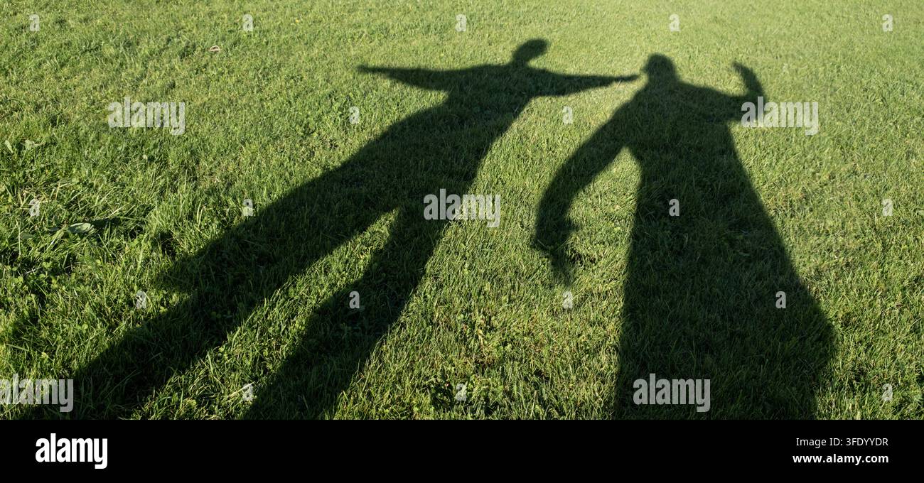 Zwei verspielte menschliche Schatten, die sich über sonnenbeschienenes grünes Gras erstrecken und ein fröhliches Outdoor-Konzept von Freundschaft, Verbindung und Freiheit schaffen. - Smartphone-aufgenommenes Stockfoto
