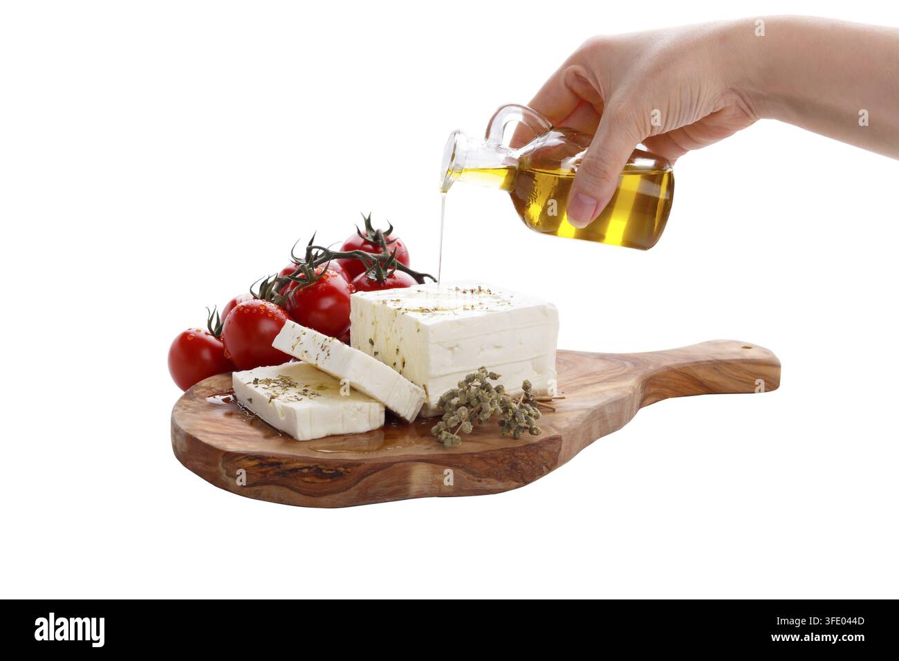 Weibliche Hand gießt Olivenöl auf griechischen Fetakäse mit Oregano und Tomaten, isoliert auf weißem Hintergrund Stockfoto