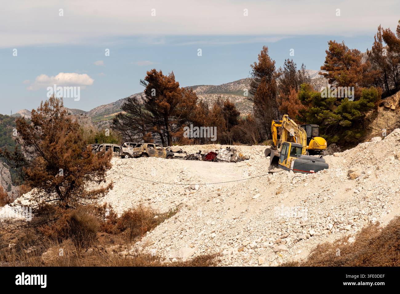 Ein gelber Bagger arbeitet neben ausgebrannten Fahrzeugen auf einem felsigen Hügel in Kroatien. Stockfoto
