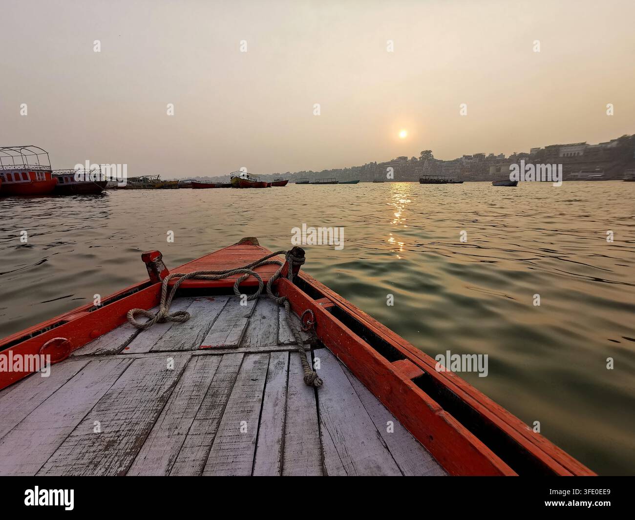 Der Bug eines Holzbootes schneidet sich durch die goldenen Gewässer des Ganges bei Sonnenuntergang in Varanasi, wobei die Ghats der Stadt und andere Boote sichtbar sind Stockfoto