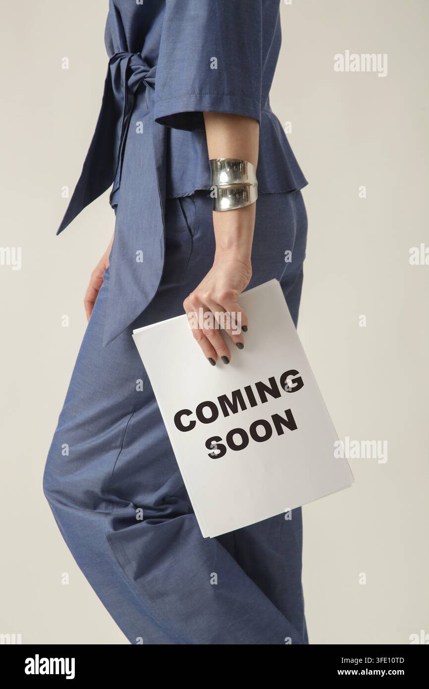 Zuschnitt einer weiblichen Model in blauem Anzug, die ein "Coming Soon"-Poster hält, kreatives Designelement für Marketingkampagne und Markenwerbung. Stockfoto