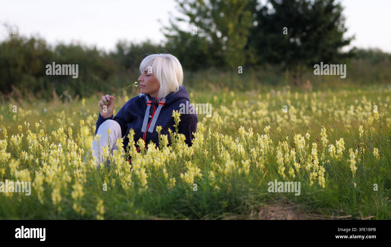 Ein friedliches Porträt einer blonden Frau in einem gemütlichen Hoodie, die in einem grünen Feld sitzt, sanft eine gelbe Wildblume mit geschlossenen Augen riecht und genießt Stockfoto
