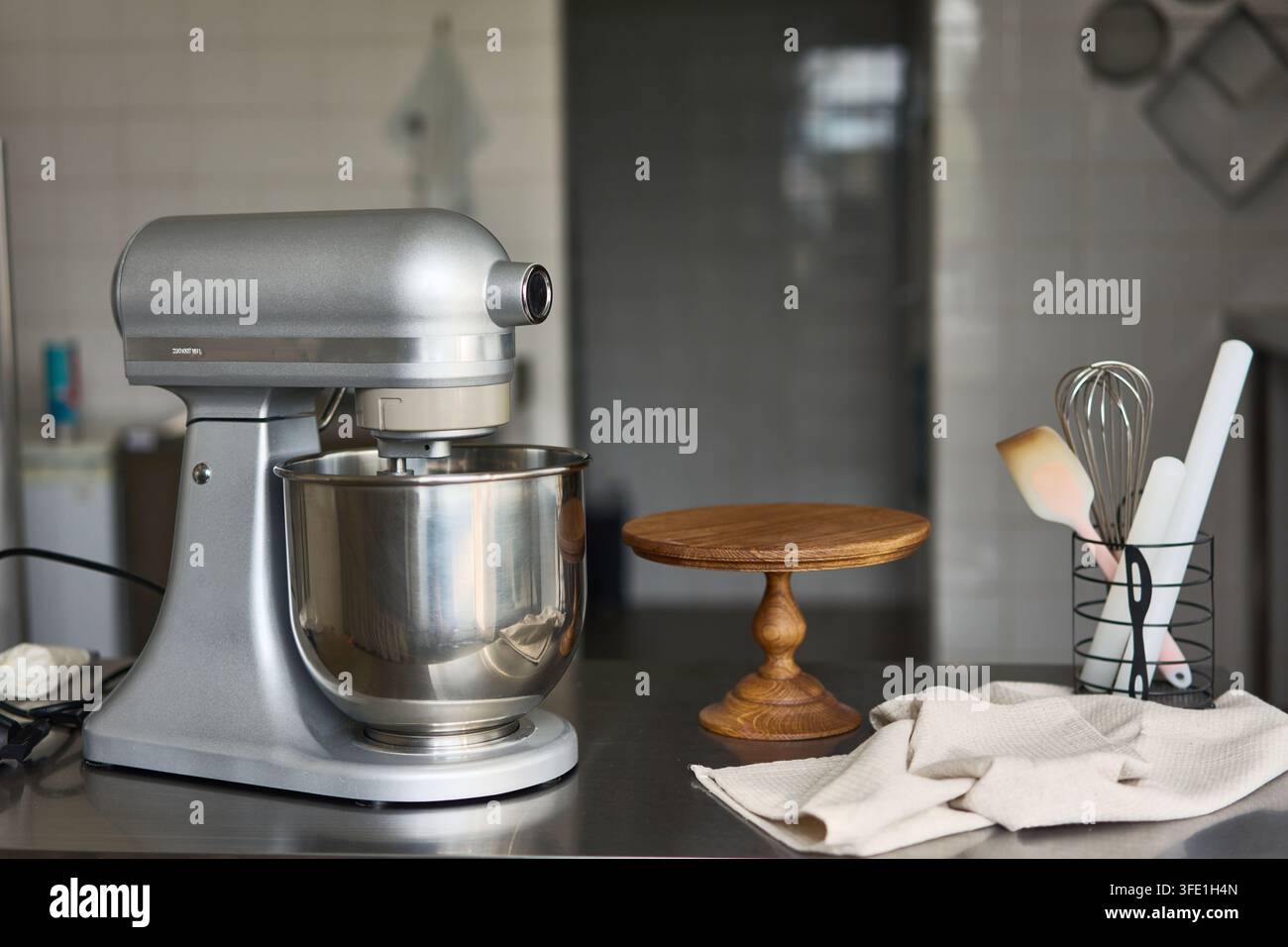 Silberner Standmixer mit Metallschüssel auf der Küchentheke neben Holz-Kuchenständer und Utensilien, mit Mixer, Stand, Silber, Metalldetail, zeigt Stockfoto