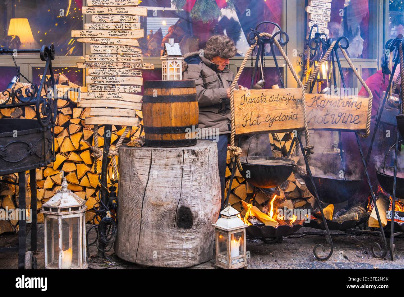 Rustikale Außenzubereitungszentren konzentrieren sich auf Eisenkessel, einen hölzernen Arbeitsblock, Feuerholz und handgeschriebene Schilder auf dem Weihnachtsmarkt in Riga für traditionelles Kochen Stockfoto