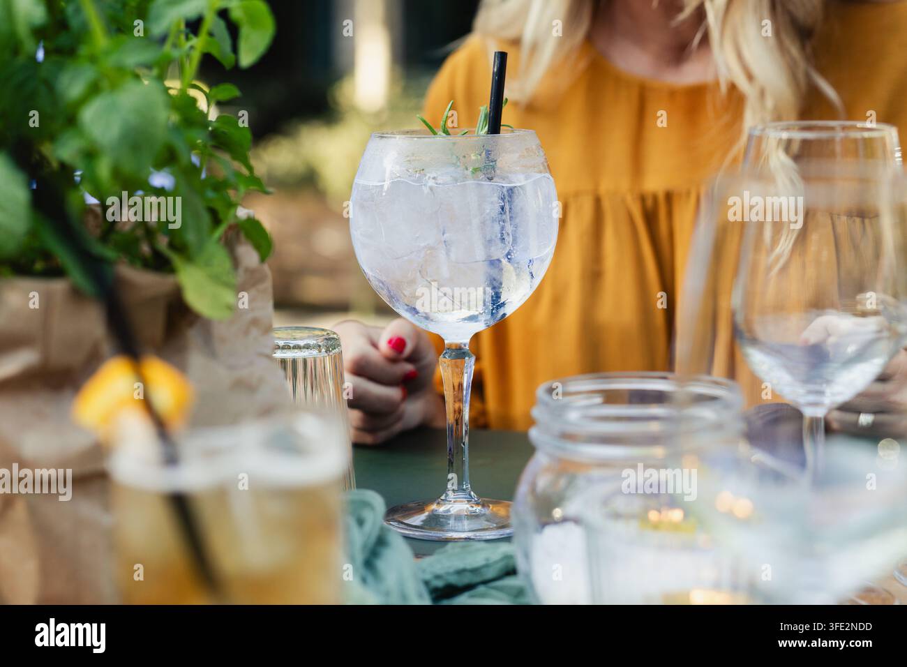 Ballonglas mit Gin Tonic mit Eis und Strohhalm, in einer boho-chicen Außeneinrichtung mit einem gedeckten Tisch. Hochzeit, Gartenparty oder Sommerabendevent Stockfoto