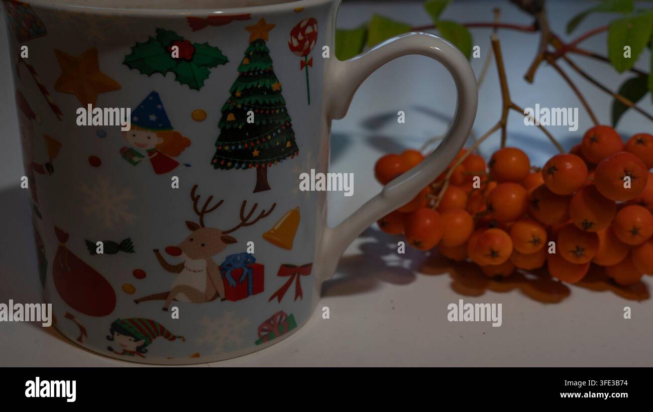 Nahaufnahme seitliche Ansicht einer festlichen weißen Keramiktasse, verziert mit bunten Weihnachtsmotiven mit Rentieren, Weihnachtsbaum und Elf, neben Orange Stockfoto