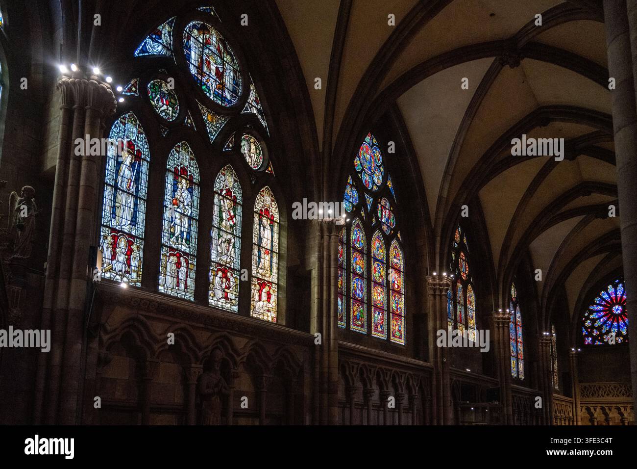 Seitenschiff einer gotischen Kirche mit beleuchteten Buntglasfenstern, Kreuzrippengewölben, Steinsäulen und dunklen architektonischen Details in Augsburg. Stockfoto