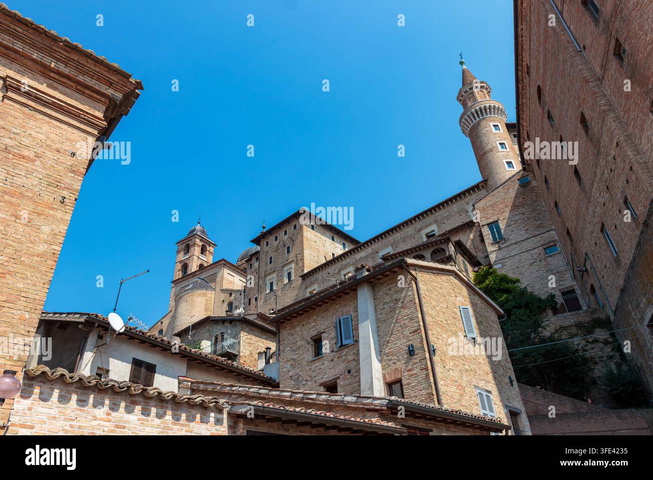 Niedrigwinkelaufnahme von historischen Backsteingebäuden und dem Palazzo Ducale-Turm unter einem klaren blauen Himmel in Urbino, Marken, Italien. Renaissance-Reiselandschaft Stockfoto
