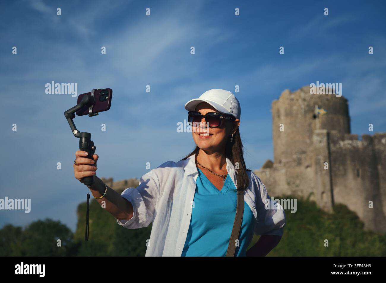 Lächelnde Reisende nimmt sich selbst mit einem Smartphone-Gimbal im Freien auf, mit einer alten Steinfortifikation und blauem Himmel im Hintergrund. Wales, Vereinigtes Königreich Stockfoto