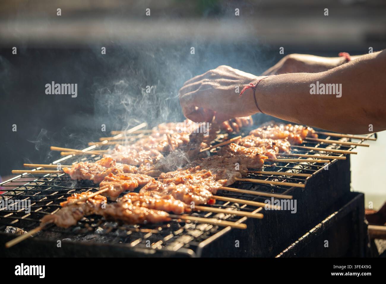 Thailändischer Mann, der Hühnerspieße über einem rauchigen Holzkohlegrill an einem Straßenessenstand im Freien grillt. Stockfoto