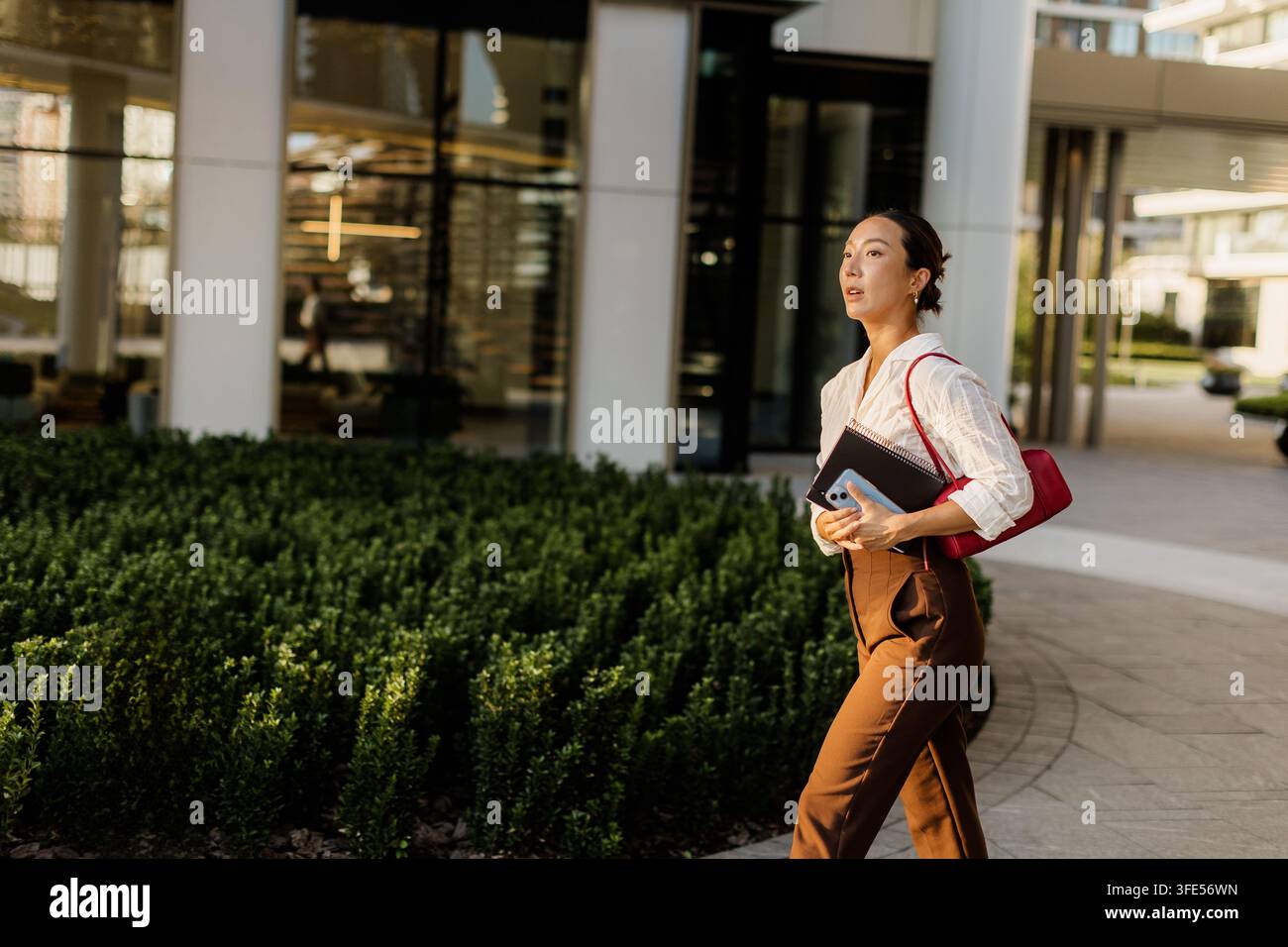 Eine koreanische Frau, stilvoll gekleidet, geht durch einen hellen und zeitgenössischen Bereich, während sie ein Tablet und ein Notizbuch hält. Sie strahlt Selbstvertrauen in ihrem Streben aus. Stockfoto