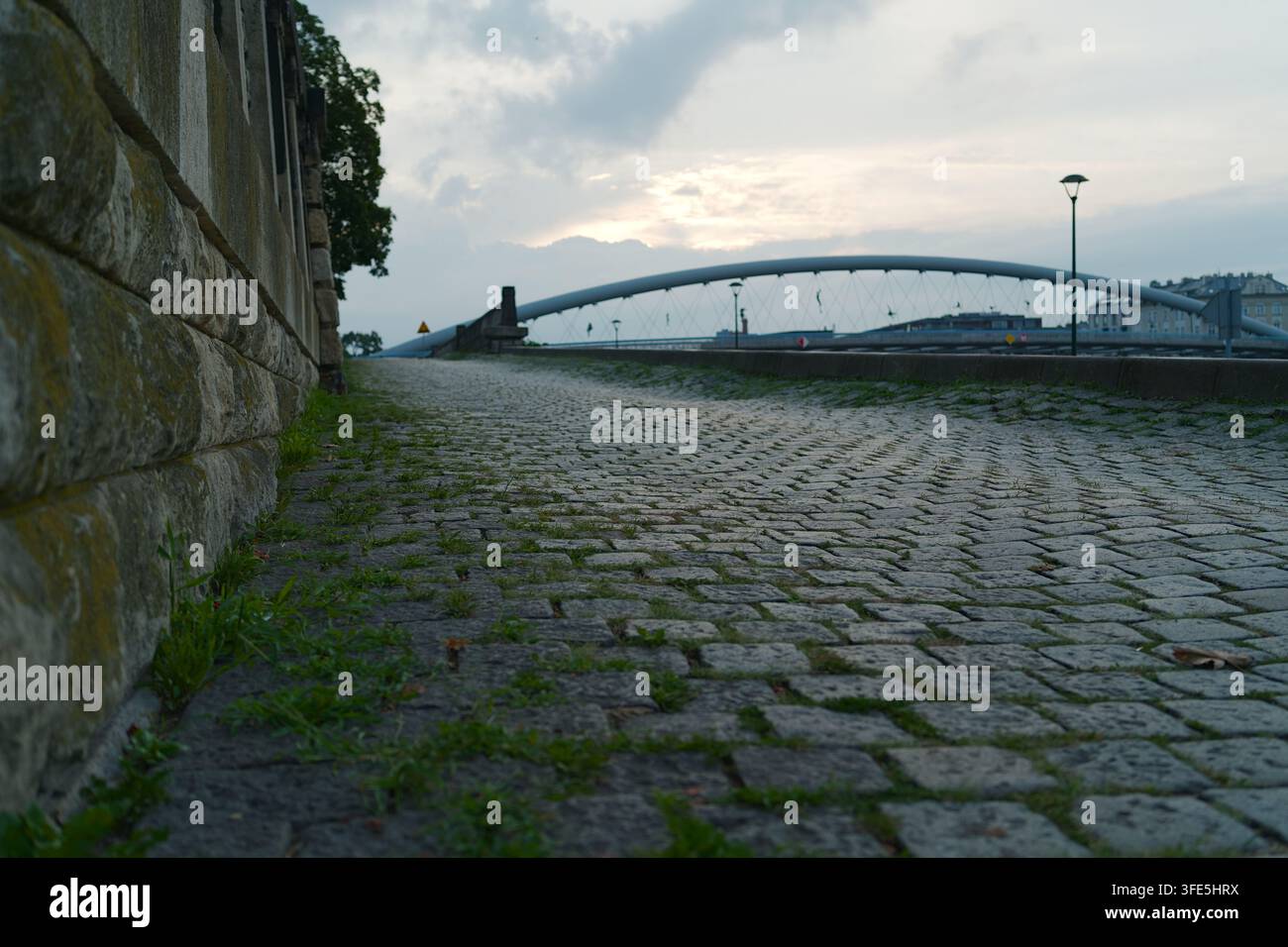 Eine niedrig angelegte Aufnahme eines alten Kopfsteinpflaster-Weges mit Gras, das zwischen den Steinen entlang einer Steinmauer wächst, der zu einer modernen Brücke führt bei d Stockfoto