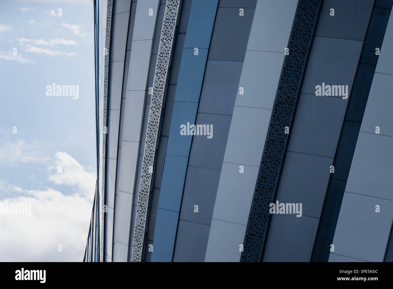 Abgewinkelte Metallverkleidungspanele fangen das Licht entlang einer modernen Gebäudefassade ein, ihre perforierten Streifen schneiden scharfe diagonale Linien gegen einen hellen Himmel Stockfoto