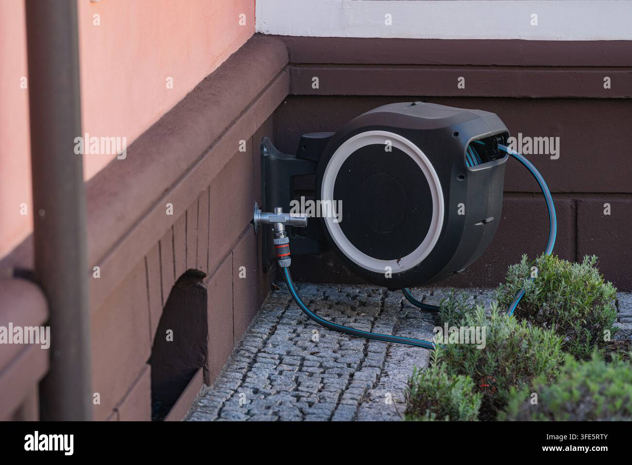 Eine Wasserschlauchhaspel ist an einer Wand neben einem Garten montiert. Die Haspel hat einen Schlauch, der an ihr befestigt ist. Der Bereich zeigt Steine und Pflanzen. Die Umgebung wirkt wie Stockfoto