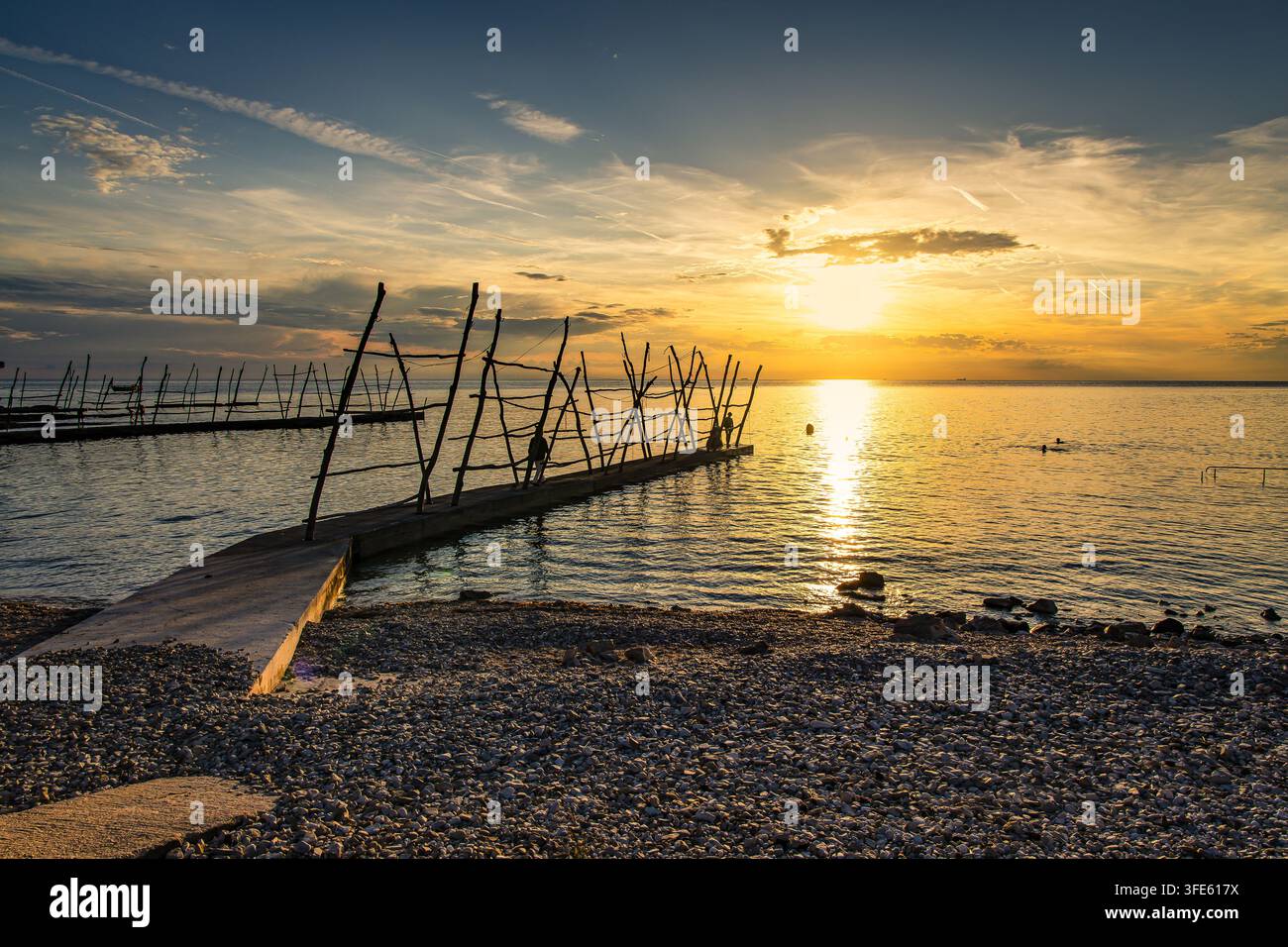 Sonnenuntergang über dem Pebble Beach Pier mit hölzernen Pfosten, die sich auf dem ruhigen Meer und im goldenen Licht spiegeln, Savudrija Stockfoto
