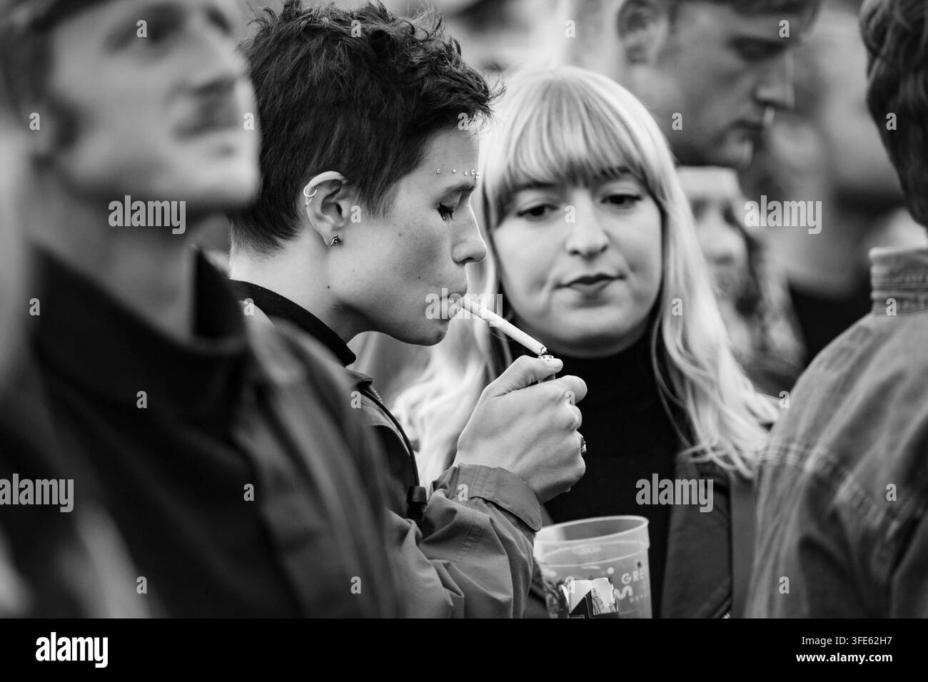 FRAU ZÜNDET EINE ZIGARETTE IN EINER FESTIVALSCHAAR AN, REPORTAGE, GREEN MAN FESTIVAL 2018, BRECON, WALES, UK, 2018: Eine junge Frau mit kurzen dunklen Haaren und Gesichts-Piercings zündet sich eine Zigarette an, während sie in einer Menge neben einer blonden Freundin steht, die roten Lippenstift trägt. Foto: Rob Watkins. INFO: Das Green Man Festival begann 2003 in Wales. Es findet jährlich am dritten Wochenende im August in den Brecon Beacons statt und feiert alternative Musik, Kunst, Literatur, Wissenschaft und familienfreundliche Kultur in einer wunderschönen Berglandschaft. Stockfoto