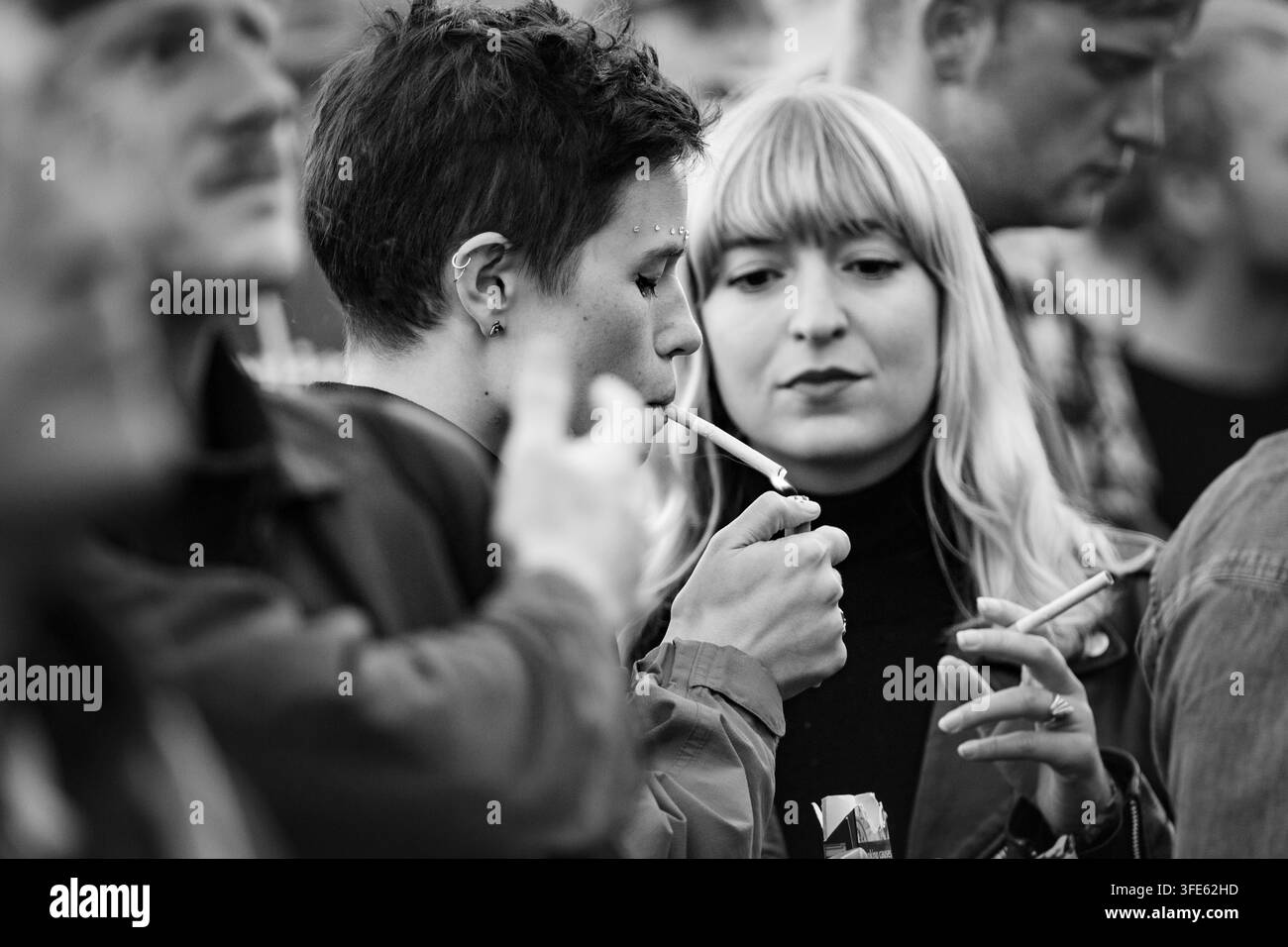 FRAU ZÜNDET EINE ZIGARETTE IN EINER FESTIVALSCHAAR AN, REPORTAGE, GREEN MAN FESTIVAL 2018, BRECON, WALES, UK, 2018: Eine junge Frau mit kurzen dunklen Haaren und Gesichts-Piercings zündet sich eine Zigarette an, während sie in einer Menge neben einer blonden Freundin steht, die roten Lippenstift trägt. Foto: Rob Watkins. INFO: Das Green Man Festival begann 2003 in Wales. Es findet jährlich am dritten Wochenende im August in den Brecon Beacons statt und feiert alternative Musik, Kunst, Literatur, Wissenschaft und familienfreundliche Kultur in einer wunderschönen Berglandschaft. Stockfoto