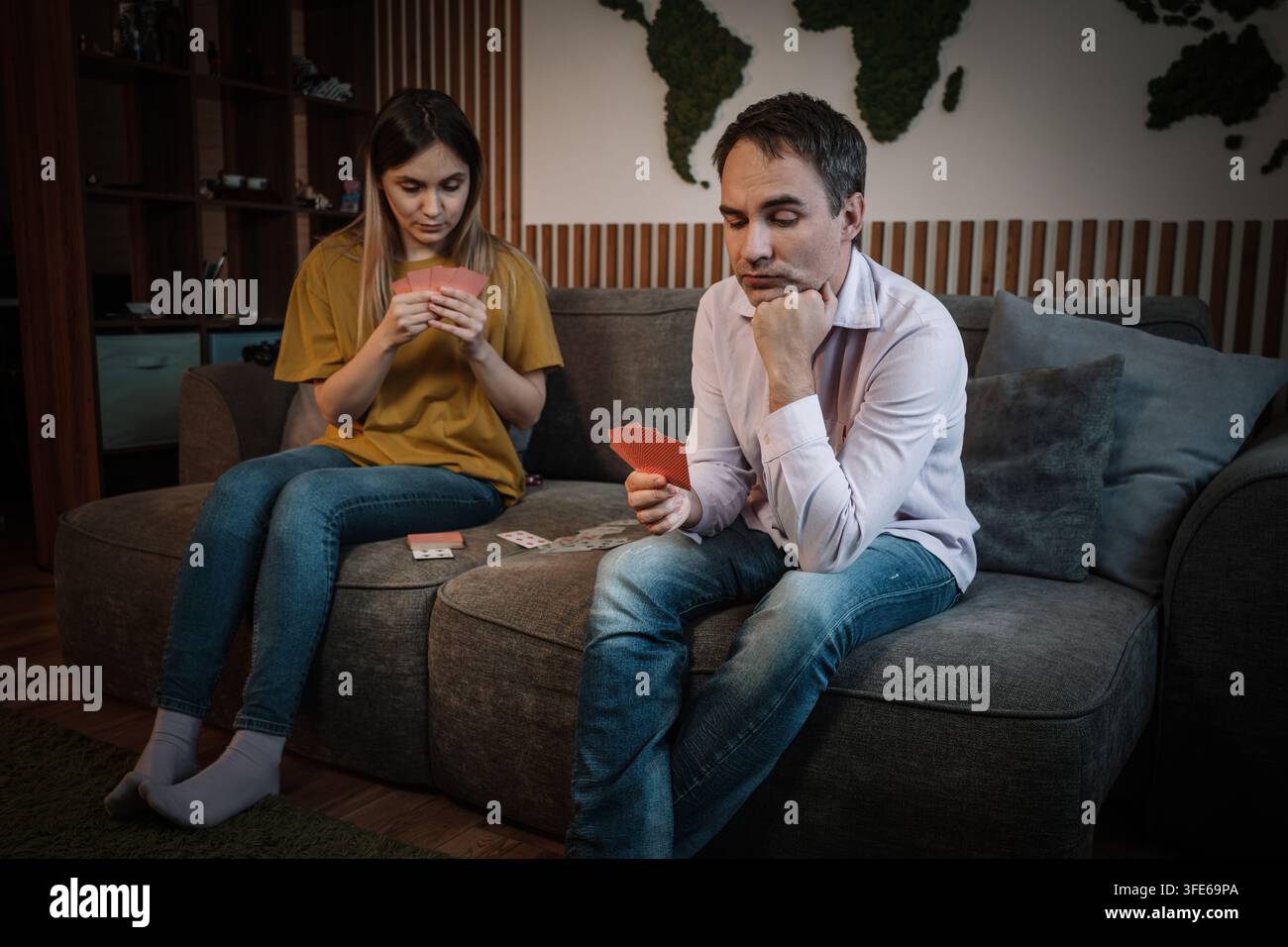 Paar spielt zu Hause Karten auf einem Sofa, beide tief auf das Spiel konzentriert, einer lächelt mit einem Hauch von Strategie. Familienunterhaltung, mentale Herausforderung, Stockfoto