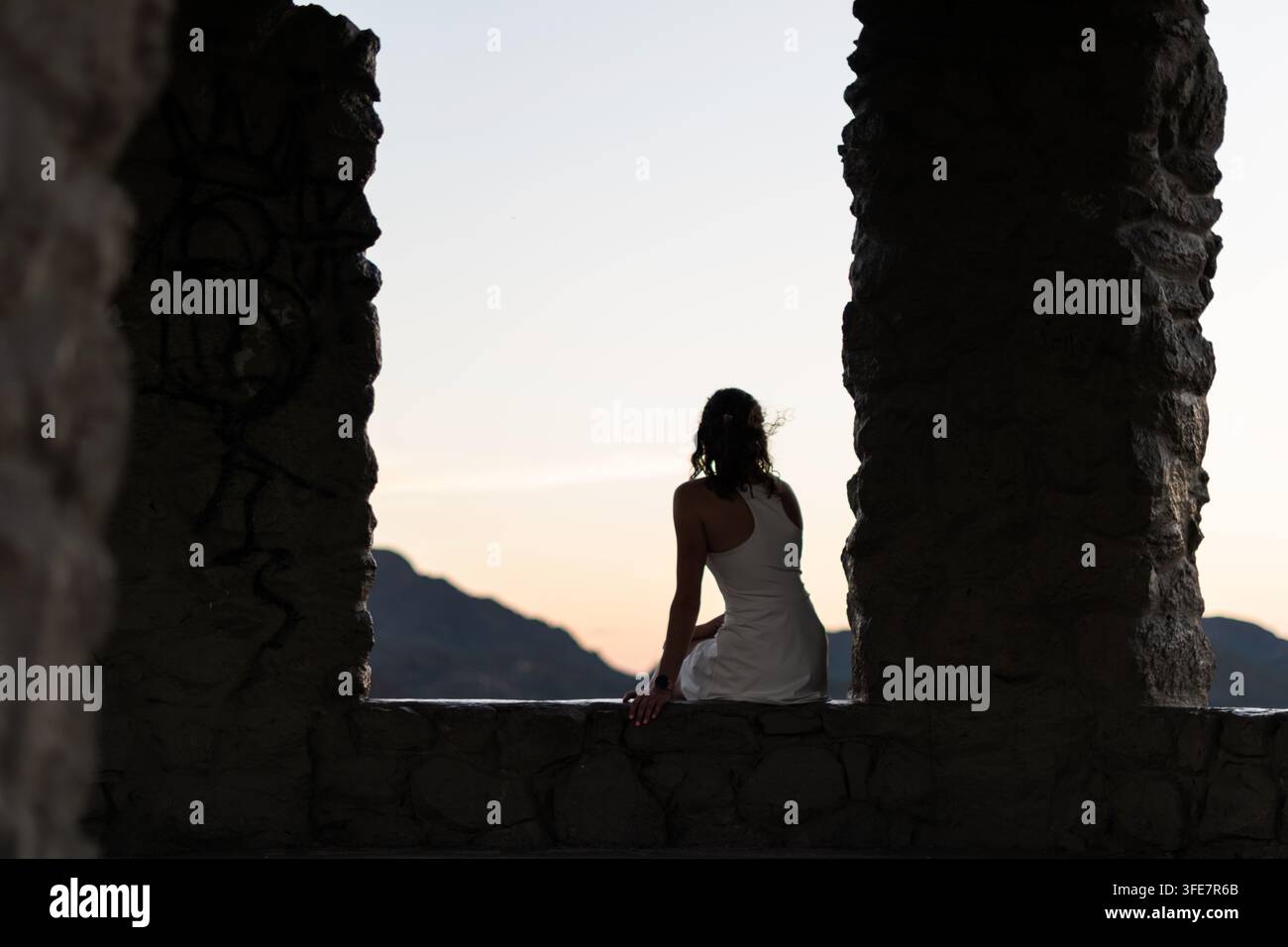 Stadtblick in der Wüste bei Sonnenuntergang - August 2022: Die Silhouette einer Frau sitzt eingerahmt zwischen zwei Steinsäulen, ferne Berge und ein leuchtender Himmel sind sichtbar Stockfoto