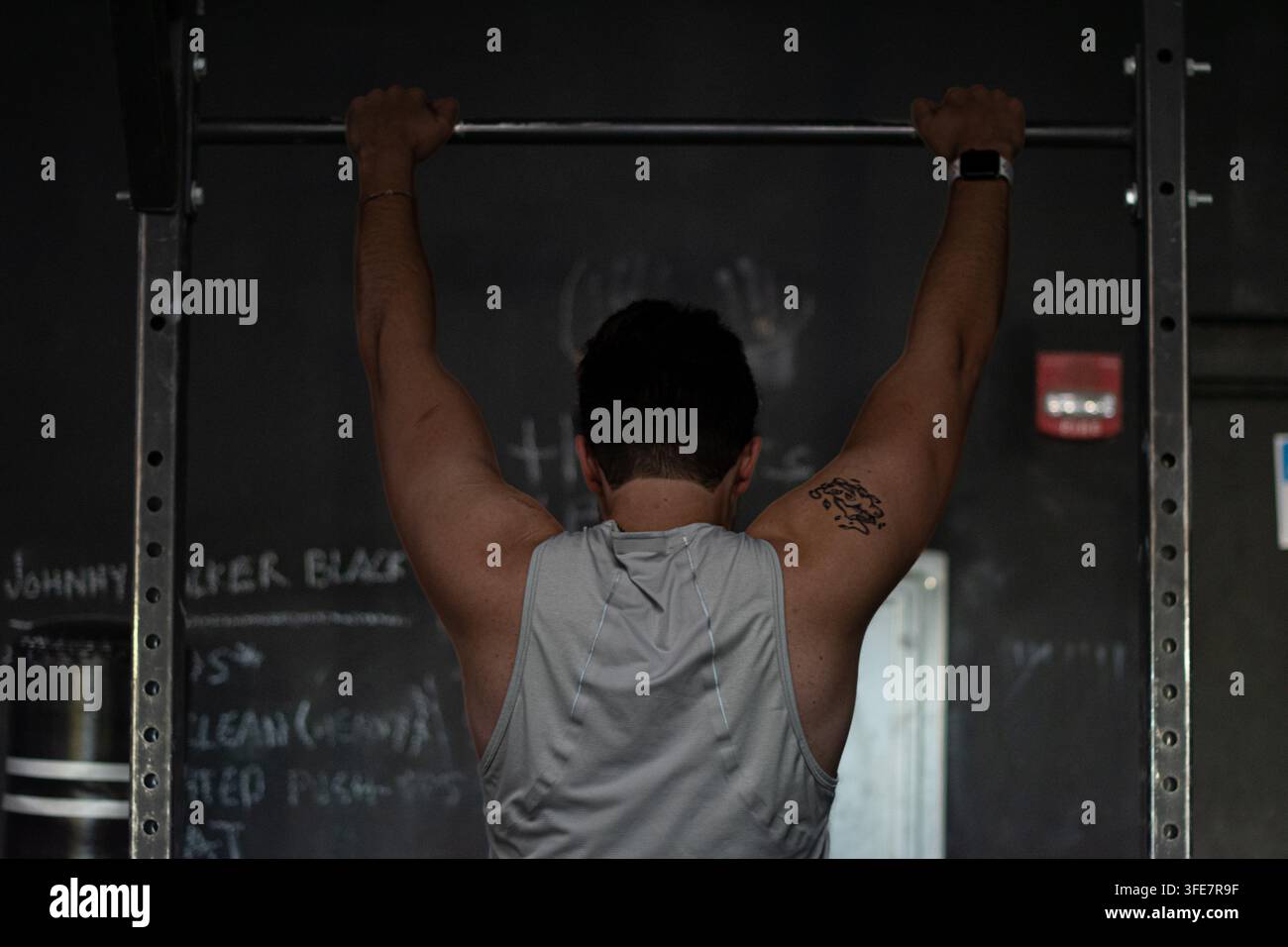 Ein muskulöser Mann führt einen Klimzug durch, von hinten betrachtet, auf einem Hantelständer in einem schwach beleuchteten Heimfitnessstudio mit Kreidetafel-Workout-Notizen an der Wand. Stockfoto