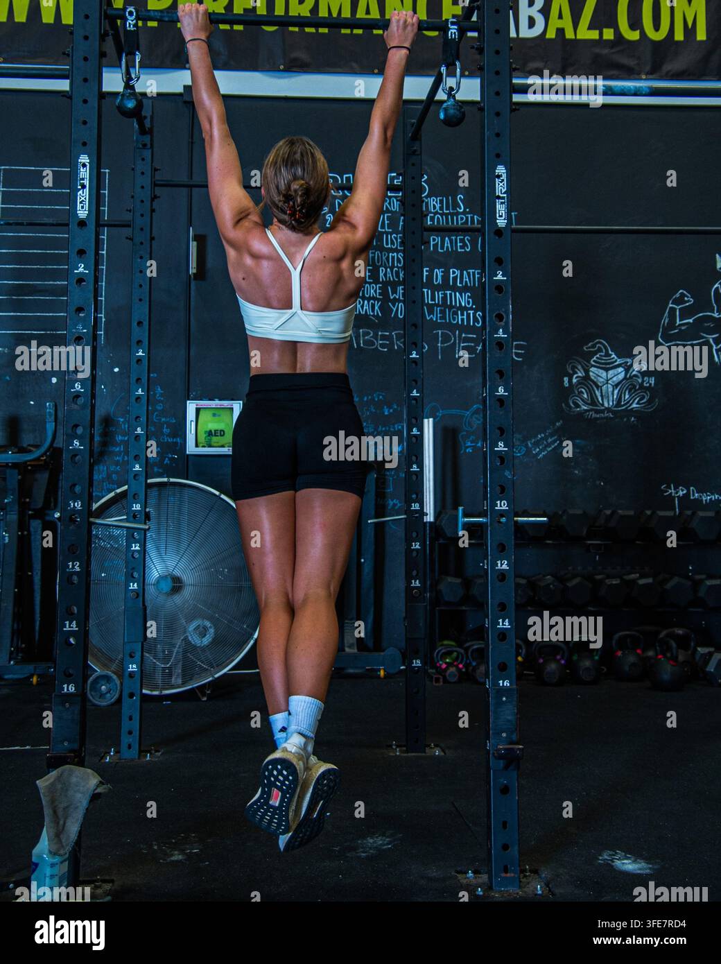 Fitnessstudio - August 2022: Eine sportliche Frau führt einen Klimzug an einer Kniebeugestange durch, die Rückenmuskeln sind angespannt, Fitnessgeräte und Kreidetafel-Schilder sind im Hintergrund sichtbar Stockfoto