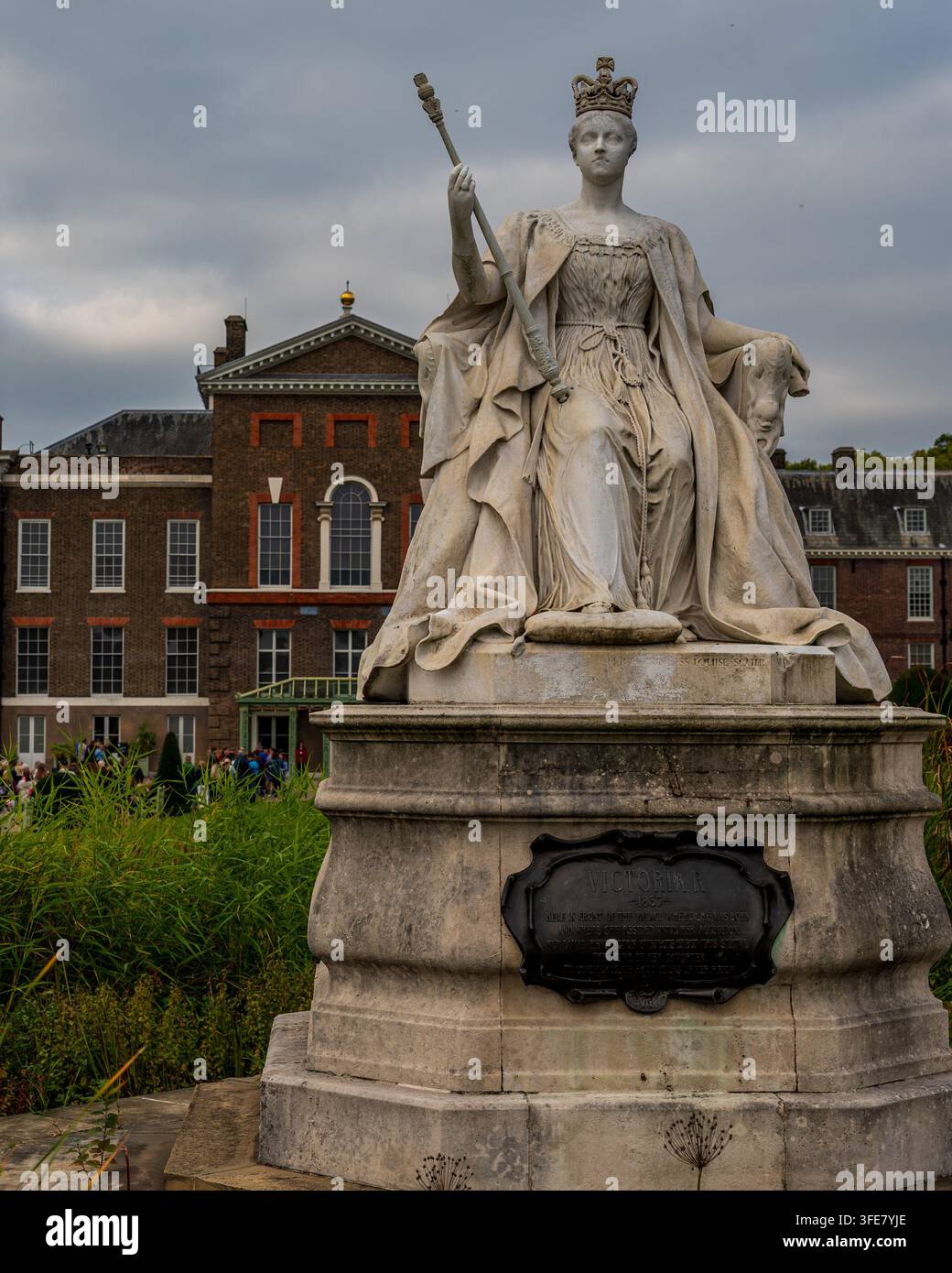 London, England – Oktober 2025: Eine Steinstatue einer sitzenden Königin Victoria, die ein Zepter hält, steht vor der rotbraunen Fassade des Kensington Palace. Stockfoto