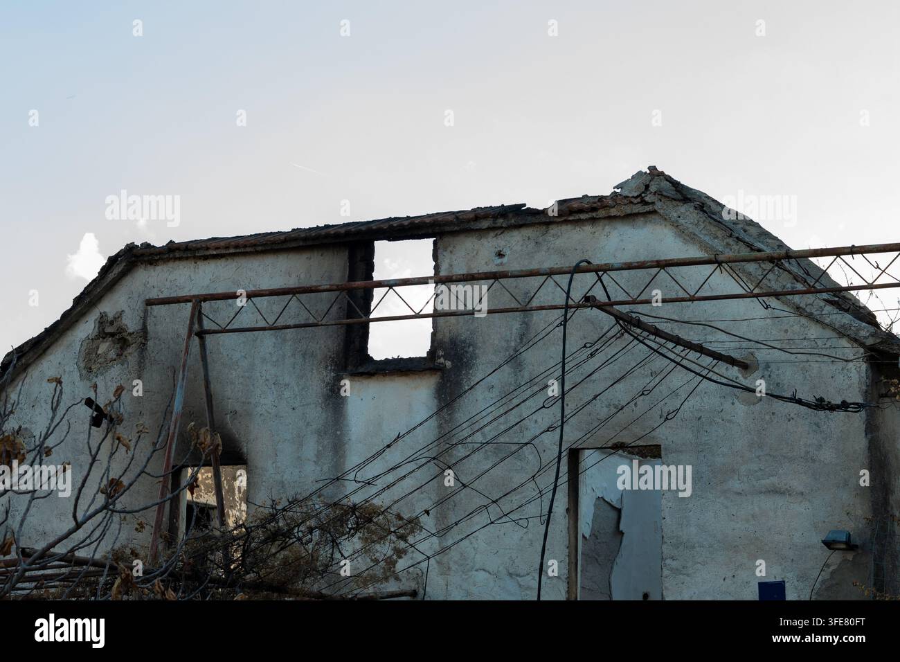 Ein beschädigtes verlassenes Haus steht unter einem blassen Himmel mit eingestürzten Dachteilen, freiliegenden Wänden und verworrenen Überkopfkabeln. Stockfoto