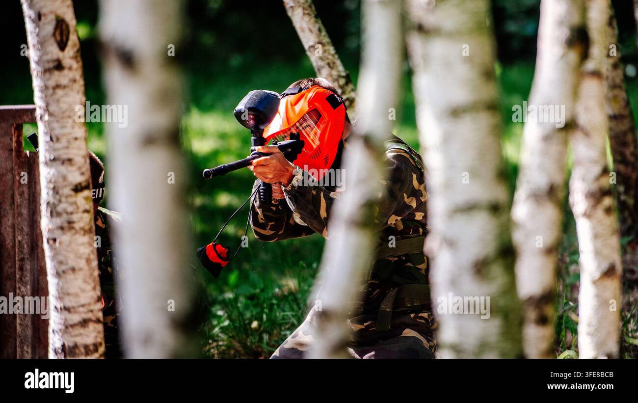 Actionaufnahme eines getarnten Spielers mit orangefarbenem Visier, der eine Paintball-Pistole durch einen Birkenhain abfeuert Stockfoto