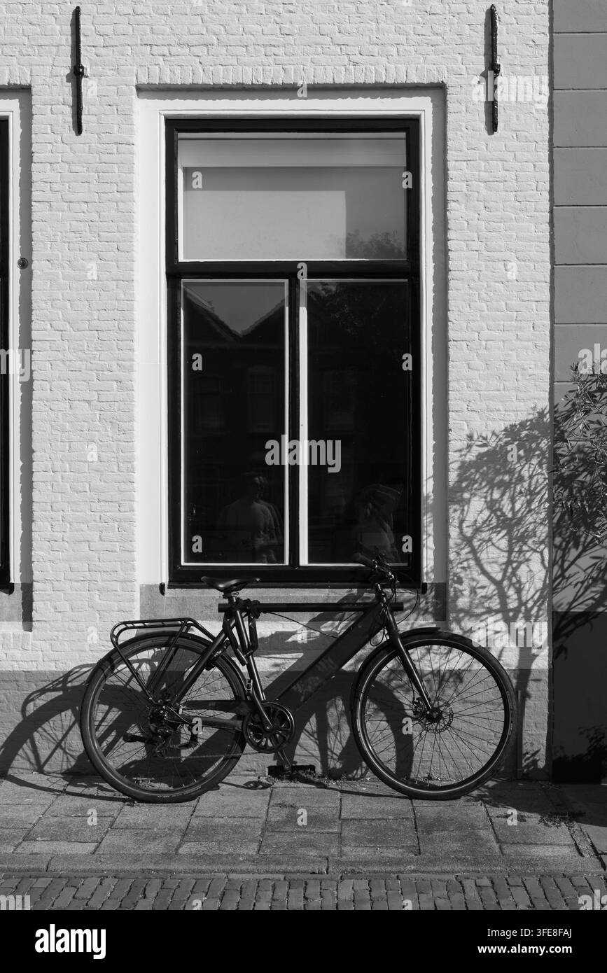 Ein Fahrrad steht an einer weiß gestrichenen Ziegelwand unter einem hohen Fenster mit starkem Sonnenlicht und grafischen Schatten an der Wand Goes Zeeland Niederlande Stockfoto
