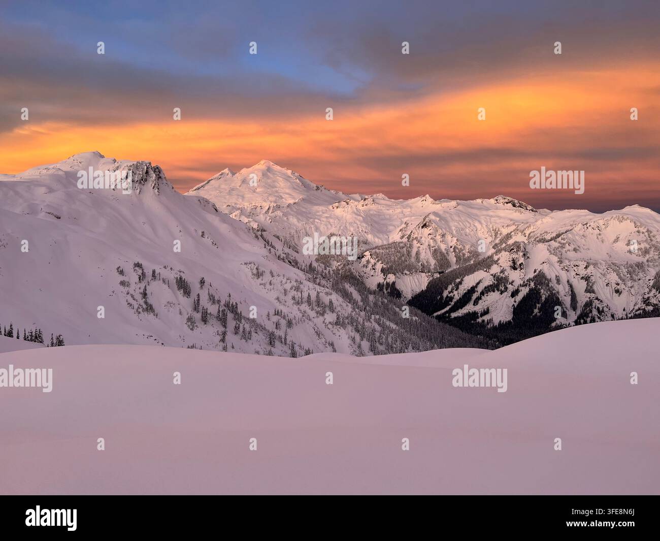 Atemberaubende winterliche Berglandschaft des Mount Baker bei Sonnenaufgang mit feurigen orangefarbenen Wolken über schneebedeckten Alpenkämmen. Stockfoto