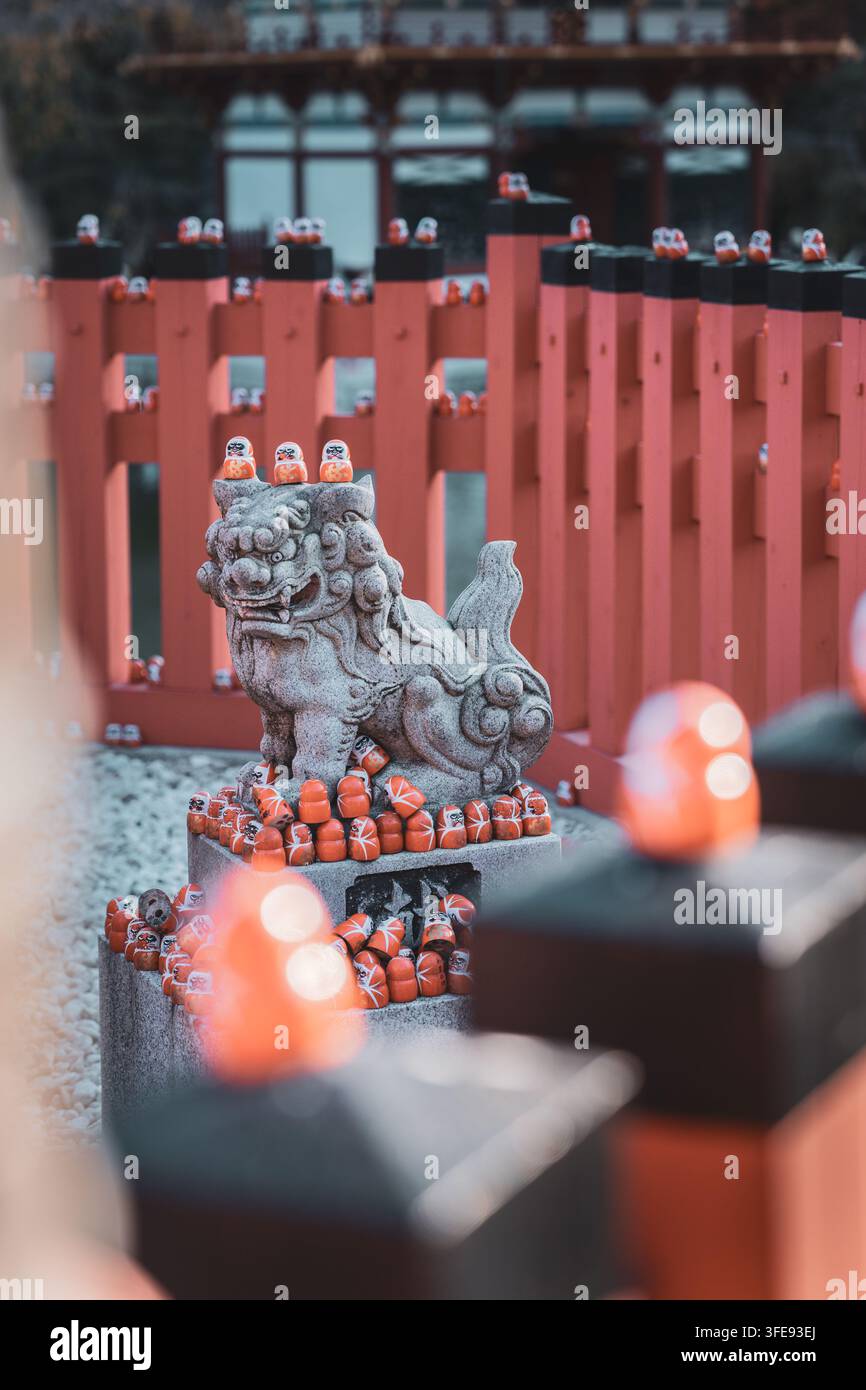 Eine geschnitzte Stein-Komainu-Wächterlöwen-Statue an einem japanischen Katsuo-ji-Schrein, oben und umgeben von kleinen roten Daruma-Puppen, mit einem roten Holzzaun Stockfoto