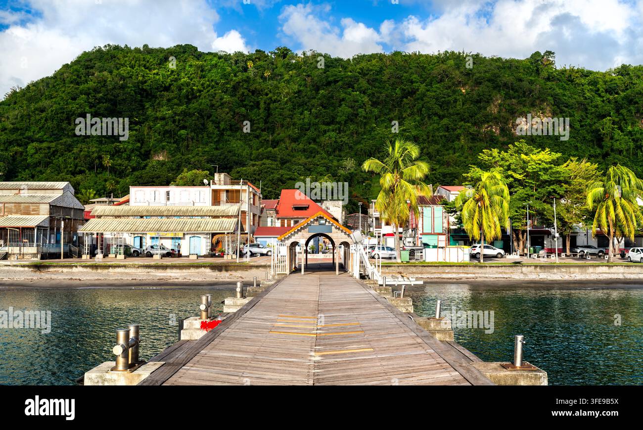 Holzsteg, der zur historischen Stadt Saint-Pierre in Martinique führt, mit üppigen grünen Hügeln, tropischen Palmen und farbenfrohen Gebäuden unter einem blauen Himmel. Stockfoto