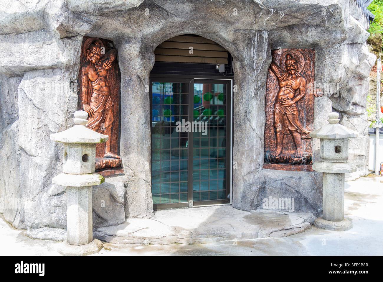 Ein kunstvoll gestaltetes Gebäudeexterior zeigt künstliche Felsformationen, kupferfarbene Figurenreliefs, dunkle Fenster und geschnitzte Steinlaternen. Stockfoto