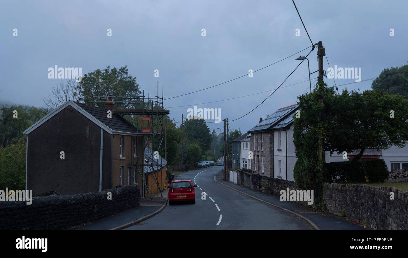 Ruhige britische Dorfstraße mit einem roten Auto, das auf einer Asphaltstraße an traditionellen Steinhäusern, Baugerüsten, Strommasten und St. Stockfoto