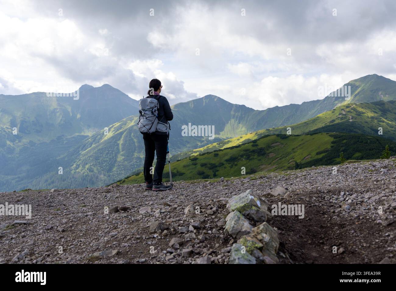 Frau steht auf einem hohen Bergpfad und betrachtet eine malerische grüne Bergkette. Stockfoto