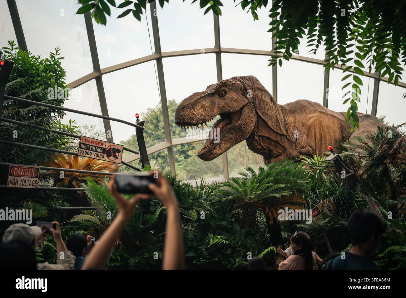 Ein großes Modell eines Tyrannosaurus-rex-Dinosauriers, umgeben von tropischen Pflanzen und Besuchern in einer Themenattraktion in Singapur. Stockfoto