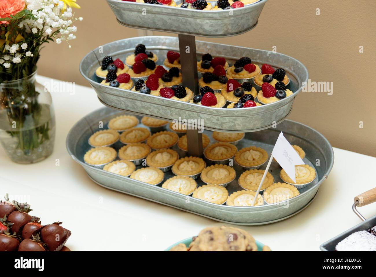 Ein dreistöckiges galvanisiertes Metalltablett, das verschiedene Mini-Fruchtschnitten und Zitronentarten auf einem Dessertbuffet-Tisch mit Blumen in der Nähe präsentiert. Stockfoto