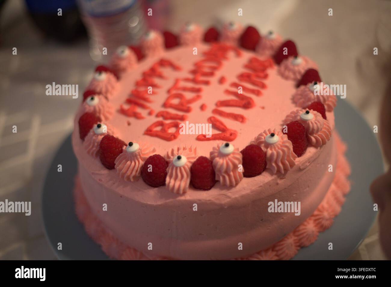 Ein runder rosa Geburtstagskuchen mit rotem Zuckerguss, auf dem die Worte „Alles Gute zum Geburtstag Vivian“ stehen, dekoriert mit frischen Himbeeren und gezogenen Zuckergussrosetten auf Stockfoto