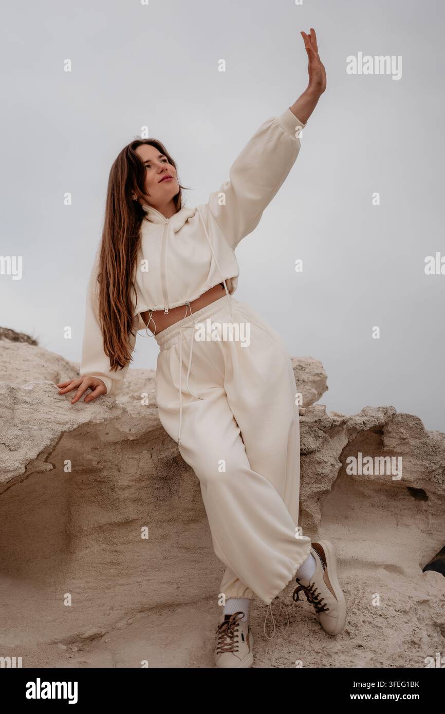 Frauen-Tracksuit-Rocks. Junge Frau in stilvoller cremefarbener Loungewear, die auf Küstenfelsen posiert mit erhobener Hand unter bewölktem Himmel Stockfoto