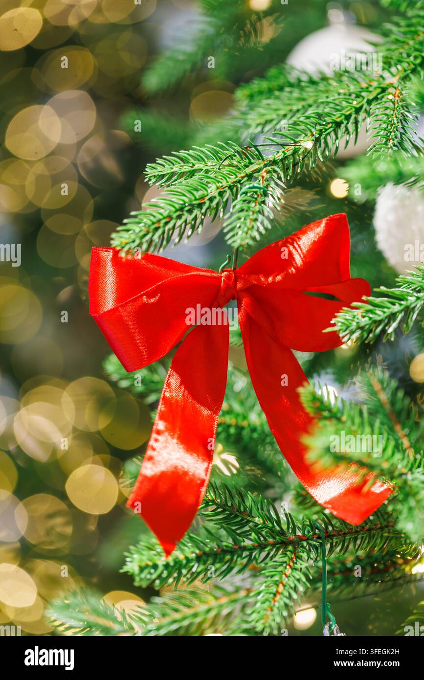 Hellrote Schleifenbänder schmücken einen grünen Weihnachtsbaum mit weißen Ornamenten und warmen goldenen Bokeh-Lichtern im Hintergrund Stockfoto