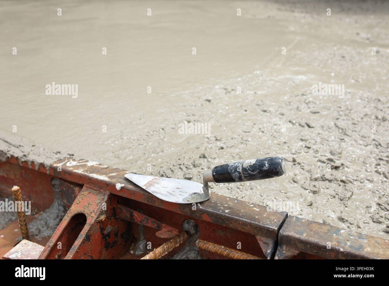 Metallkelle ruht auf Stahl Schalungskante neben glatt fertiggestelltem Betonoberfläche, Werkzeuge markieren Pause im Bodenausgleichsprozess bei aktiver Baustelle Stockfoto