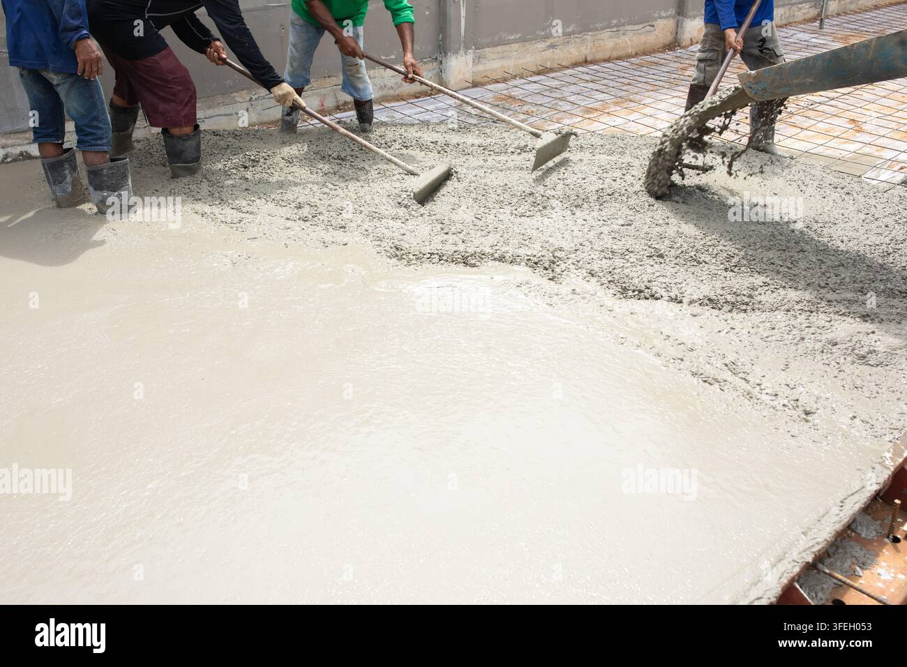 Bauarbeiter verteilen nassen Beton, der aus dem Mischtrichter gegossen wird, über den Fundamentbereich, Werkzeuge in der Hand, um das frische Material zu glätten, während graue Flüssigkeit fließt Stockfoto